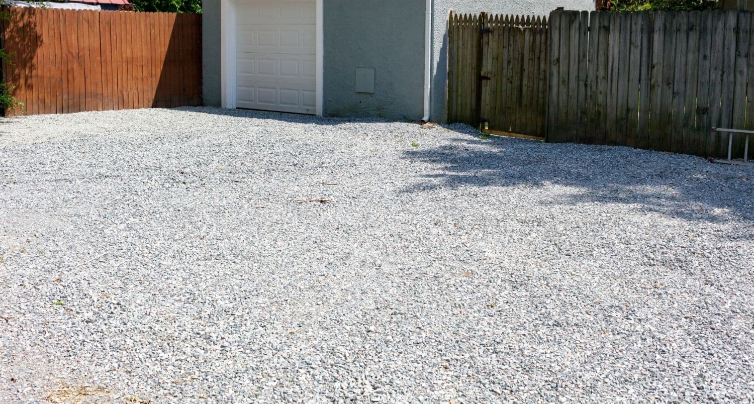 A gray crushed stone driveway and parking area running up to a garage between timber fences