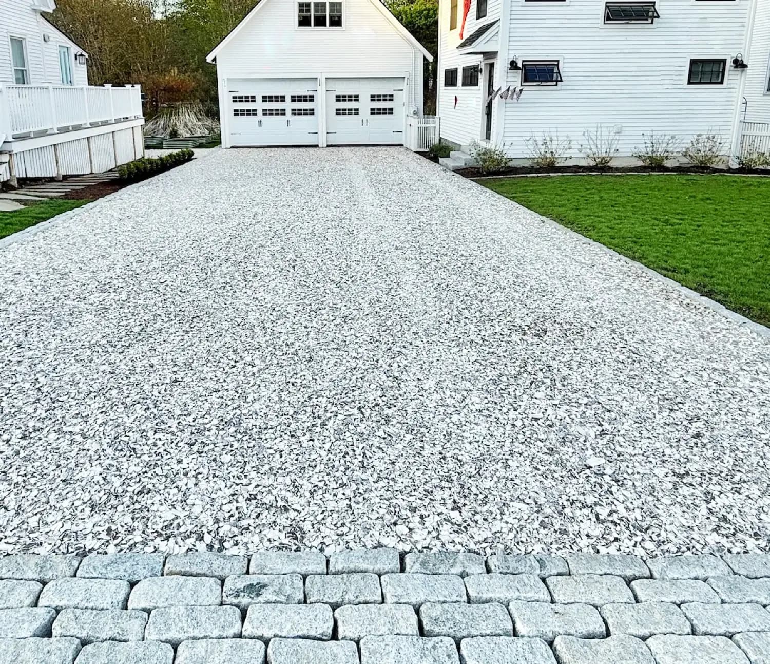 A crushed shell driveway running between two white Cape Cod houses to a garage, finished at the road with a granite cobble apron